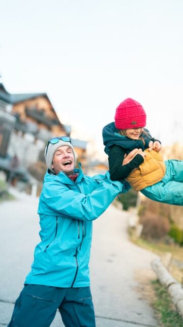 Père enthousiaste portant son enfant en tenue d'hiver dans le village de Vaujany